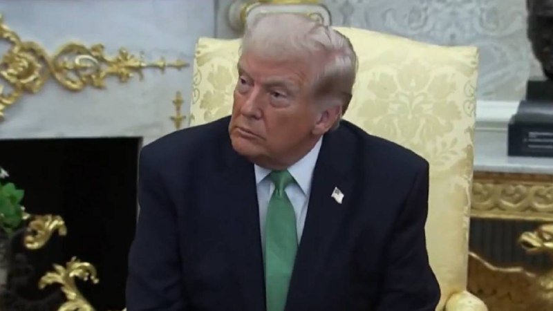 A man with blonde hair wearing a dark suit, white shirt, and green tie sits in an ornate gold-upholstered chair in a formal room.