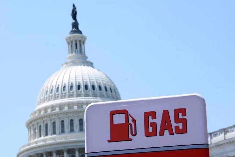 A white "GAS" station sign with a red fuel pump icon stands in the foreground, with the white domed U.S. Capitol building visible against a clear blue sky behind it.