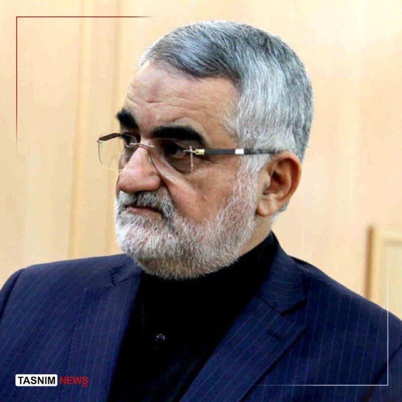 A gray-haired, bearded man wearing glasses and a dark pinstriped suit looks off-camera against a beige interior wall, with a "Tasnim News" watermark visible.