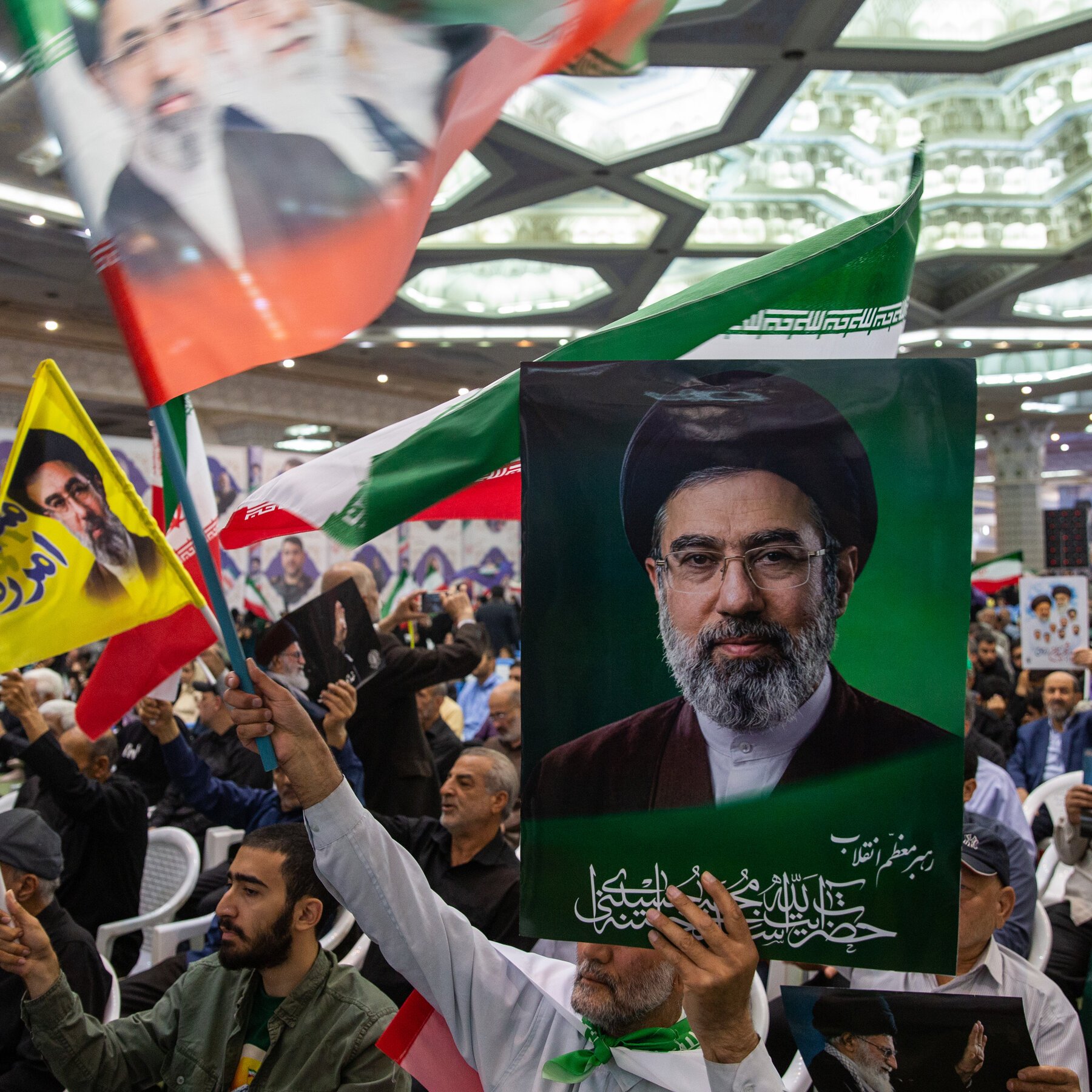 A crowd waves Iranian flags and holds up a portrait of a bearded man in clerical attire during a gathering inside a large hall.