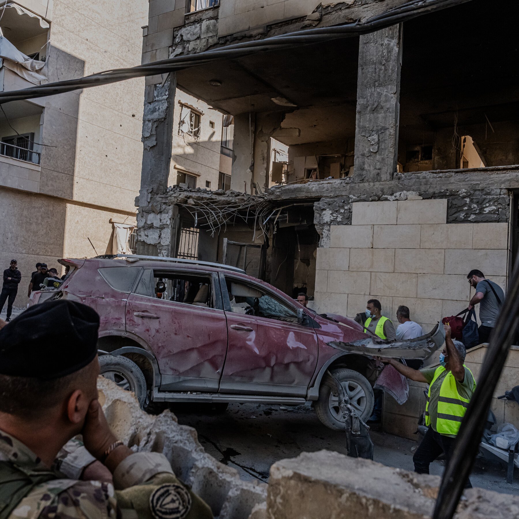 Workers in high-visibility vests clear debris and move a damaged red SUV near a heavily destroyed multi-story building with exposed rebar and shattered concrete.