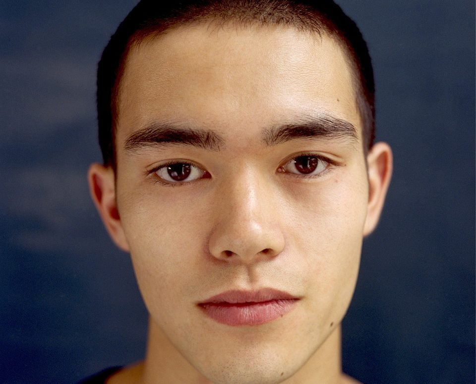 A close-up portrait of a young man with short dark hair, looking directly at the camera against a gray background.