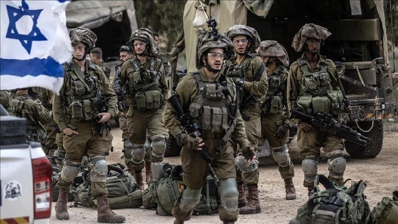 Soldiers in olive-green uniforms, helmets, and body armor stand with rifles near a vehicle, with an Israeli flag visible on the left.