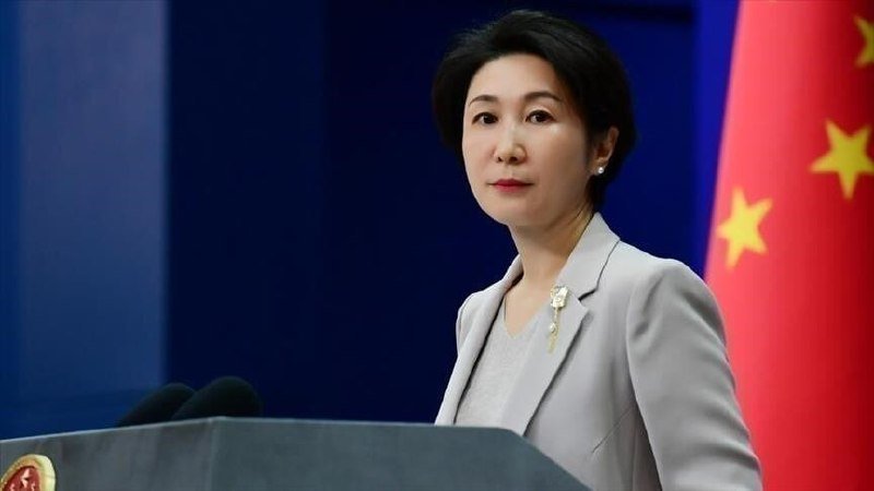 A woman in a gray blazer stands at a podium with microphones, with a Chinese flag visible behind her against a blue backdrop.