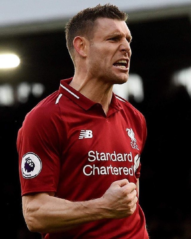 A short-haired soccer player in a red Liverpool FC jersey with "Standard Chartered" and Premier League patches clenches his fist while shouting during a match.