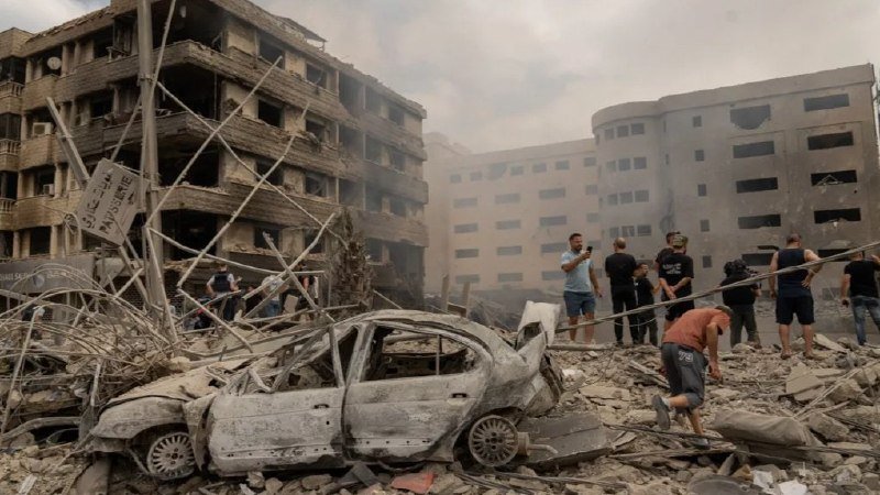 A burned-out car sits amid rubble in front of damaged multi-story buildings as several people stand surveying the debris-strewn site.