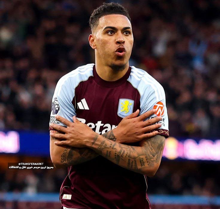 A tattooed football player in a claret and light blue Aston Villa jersey crosses his arms in a celebration pose before a blurred stadium crowd.
