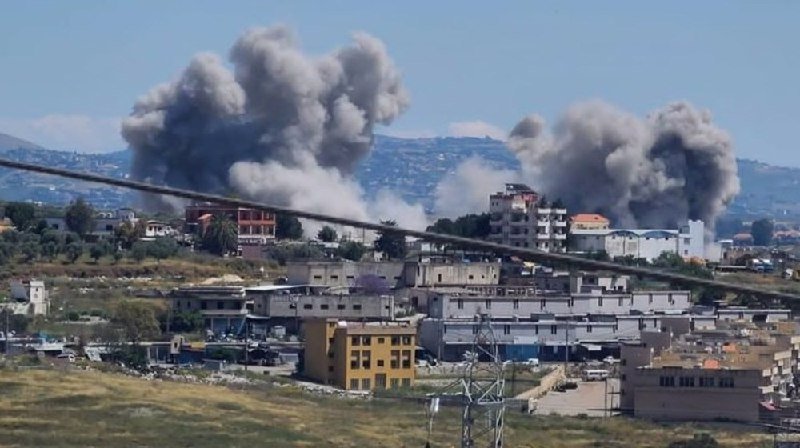 Large plumes of gray smoke rise above a densely built urban area with multi-story buildings, with hills visible in the background.