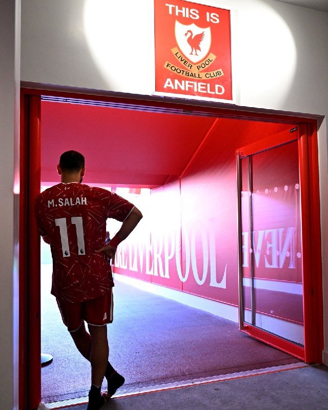 A player wearing a red "M. SALAH 11" jersey walks through a red-framed stadium tunnel beneath a "THIS IS ANFIELD" sign toward a green pitch.
