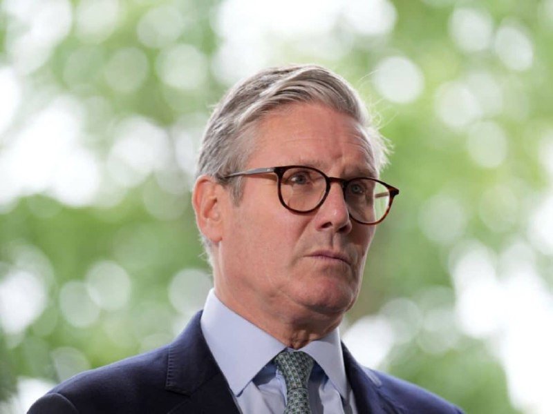 A gray-haired man wearing glasses, a dark suit, light blue shirt, and patterned tie stands outdoors against a blurred green background.