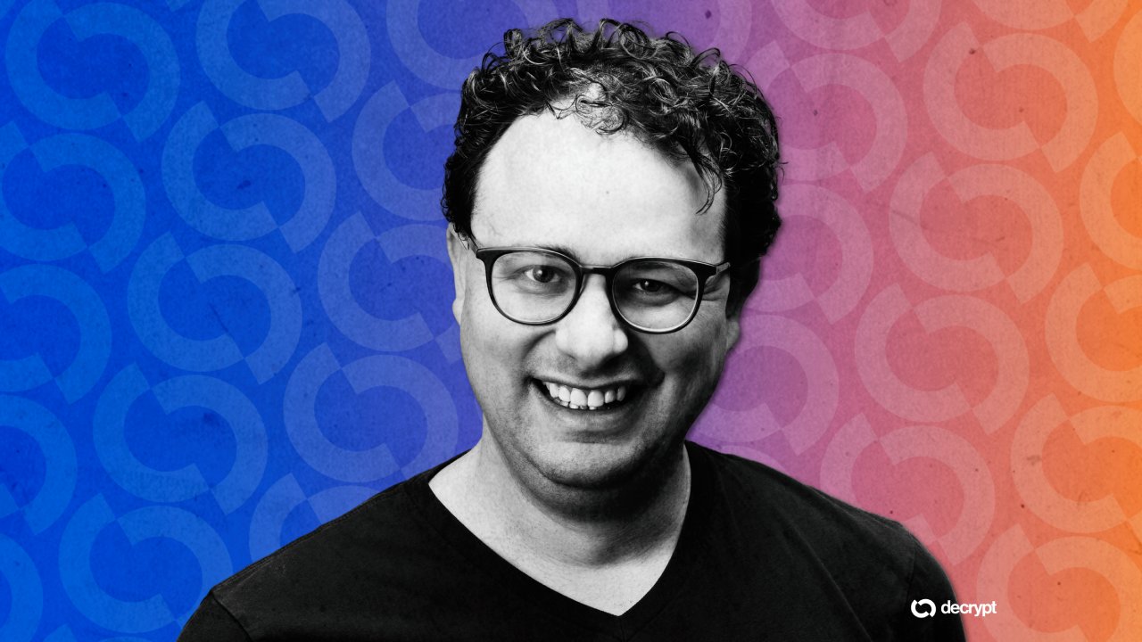 A black-and-white photo of a smiling, curly-haired man wearing glasses and a dark t-shirt against a blue-to-orange gradient background with a "decrypt" logo.