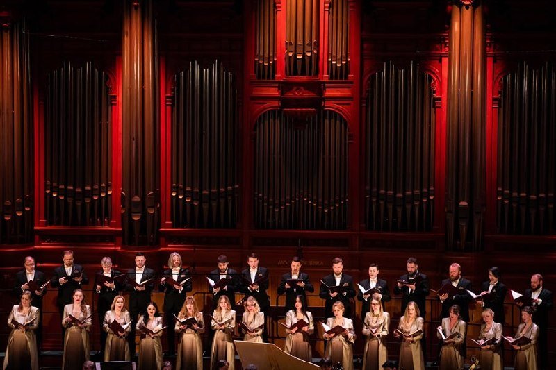 A choir in formal attire performs on stage holding folders of sheet music, set against an ornate pipe organ illuminated in red light.