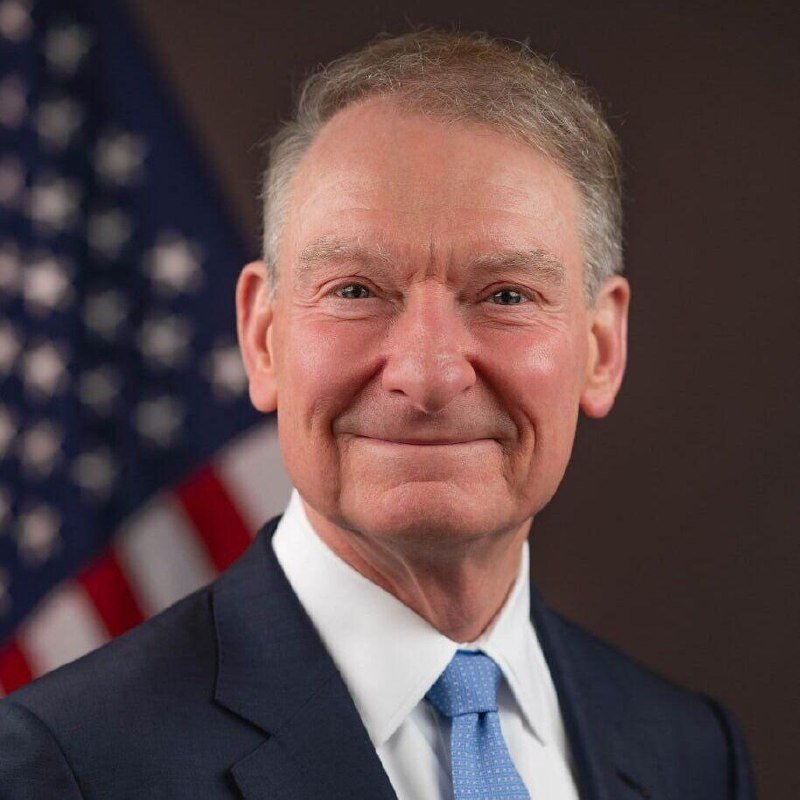 An older man with gray hair, wearing a navy suit, white shirt, and blue tie, smiles at the camera with an American flag visible behind him.