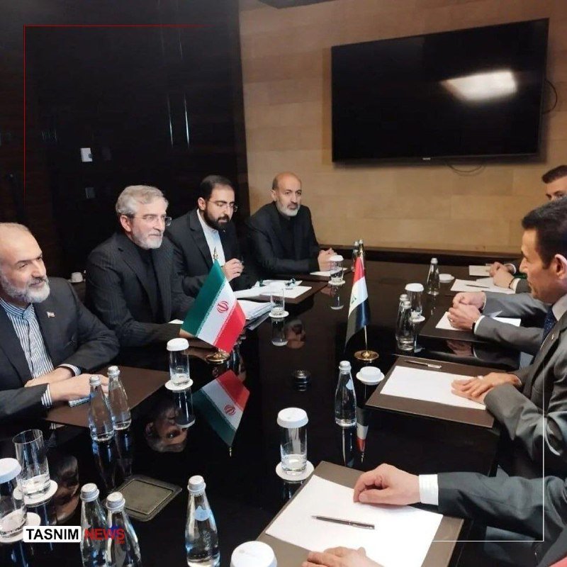 Several men in suits sit around a conference table with water bottles, papers, and small Iranian and Iraqi flags, conducting a meeting.