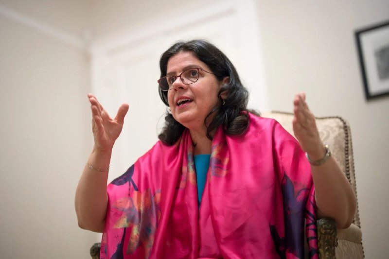 A bespectacled woman in a pink patterned scarf gestures with both hands raised while speaking indoors, seated in an upholstered chair.