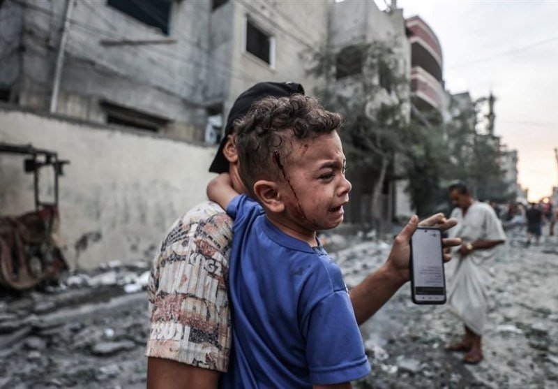 A bloodied, crying child in a blue shirt is held by a man in a graffiti-patterned shirt, who holds up a smartphone displaying text, amid a street strewn with rubble and damaged buildings.