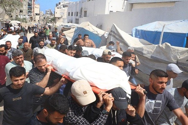 A crowd of men carry a body wrapped in a white shroud on a stretcher through a street lined with white and blue tarpaulin shelters amid damaged buildings.