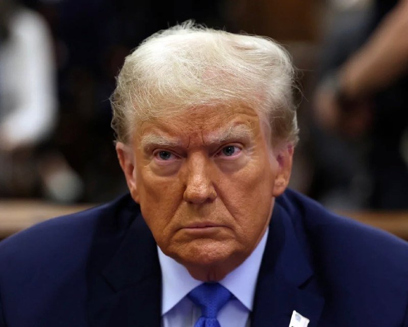 A man with blonde hair wearing a dark blue suit, light blue shirt, and blue tie looks forward with a serious expression.