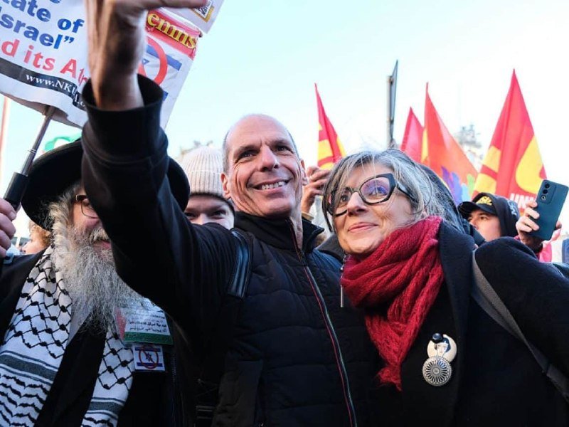 A smiling man and woman pose together at an outdoor gathering, surrounded by people holding red flags, placards, and a partially visible sign referencing "Israel" and "its A...," with a keffiyeh-patterned scarf visible on a nearby attendee.