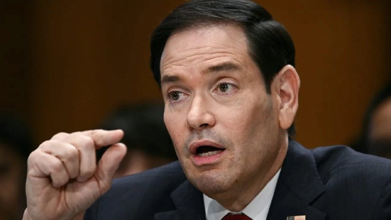A man in a dark suit with an American flag pin gestures with a pinched hand while speaking.