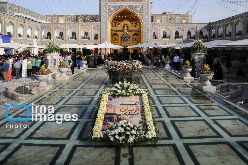 A floral arrangement with Persian script sits in a tiled courtyard of an ornate shrine, surrounded by crowds gathered beneath a grand golden iwan.