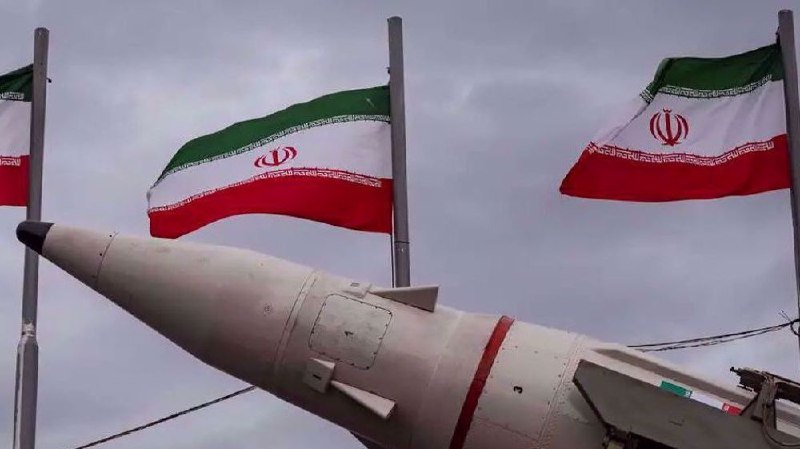 A large white missile is displayed outdoors beneath three Iranian flags waving against a gray sky.