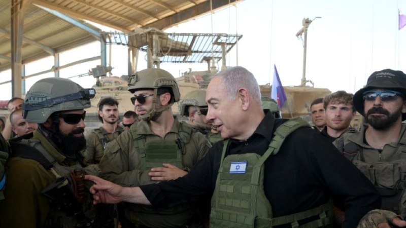 A man in a black shirt and tactical vest speaks with soldiers in helmets and combat gear, with armored vehicles visible under a large open hangar.