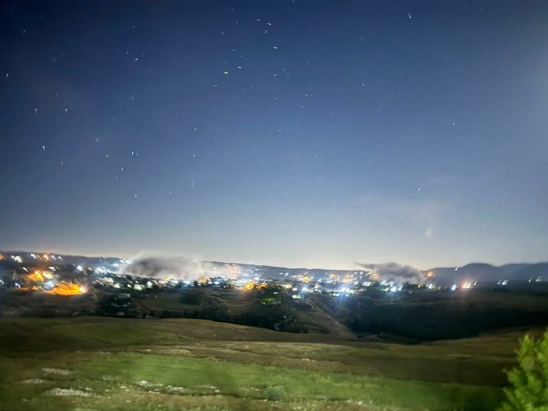 A nighttime long-exposure photo shows a starry sky above a distant, illuminated valley town, with dark grassy hills in the foreground.