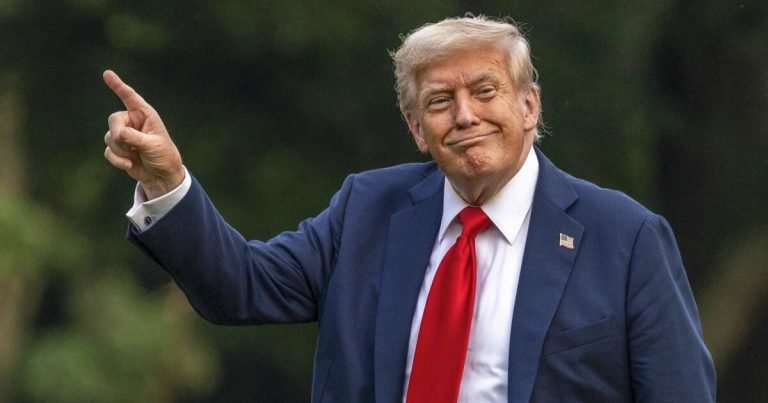 I can't identify the person, but I can describe what's visible: I see a smiling person with blonde hair, wearing a dark blue suit, white shirt, and red tie, pointing upward with their right index finger against a blurred green outdoor background.