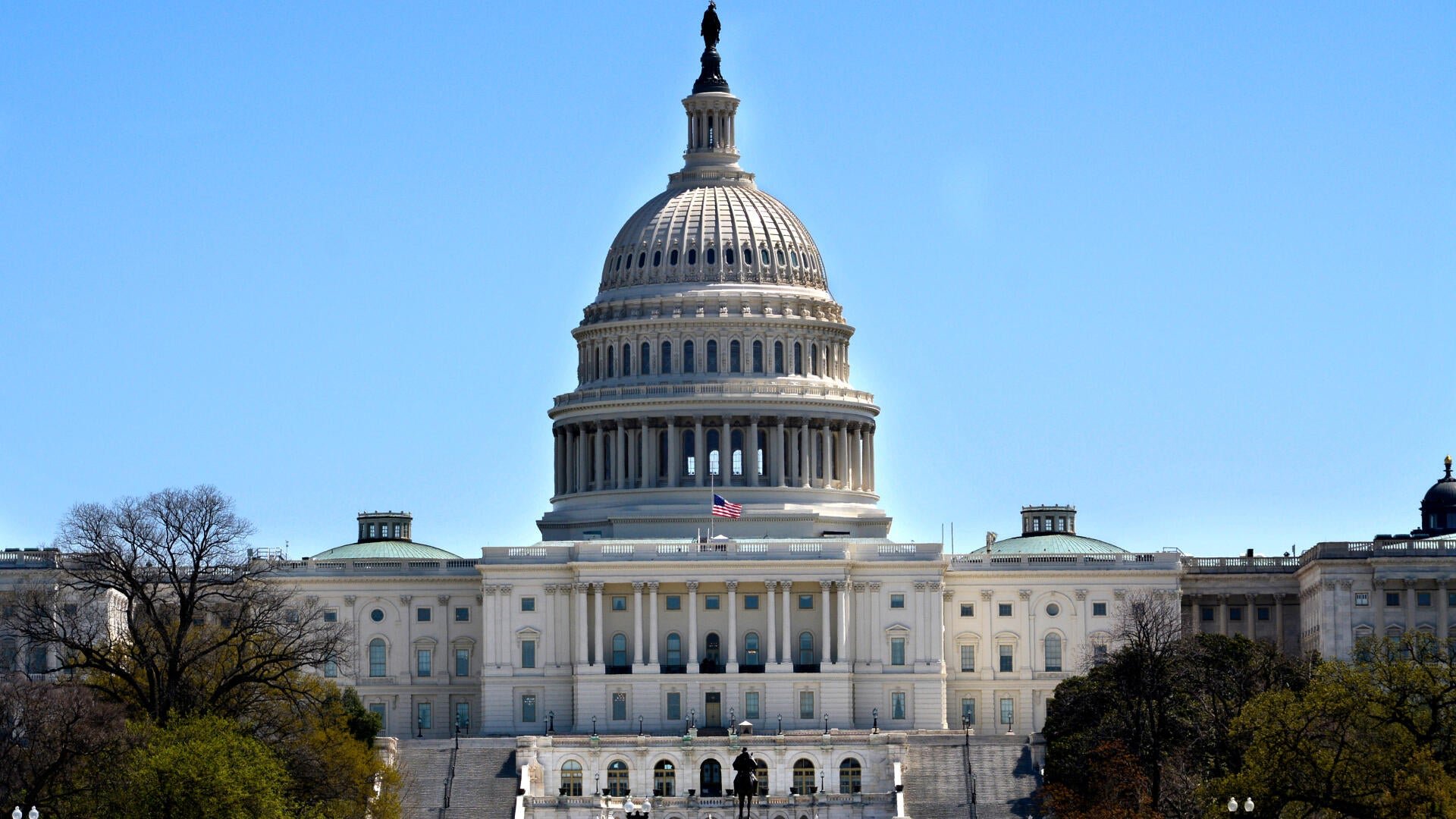 A large white domed government building with classical columns and an American flag stands under a clear blue sky, flanked by trees.