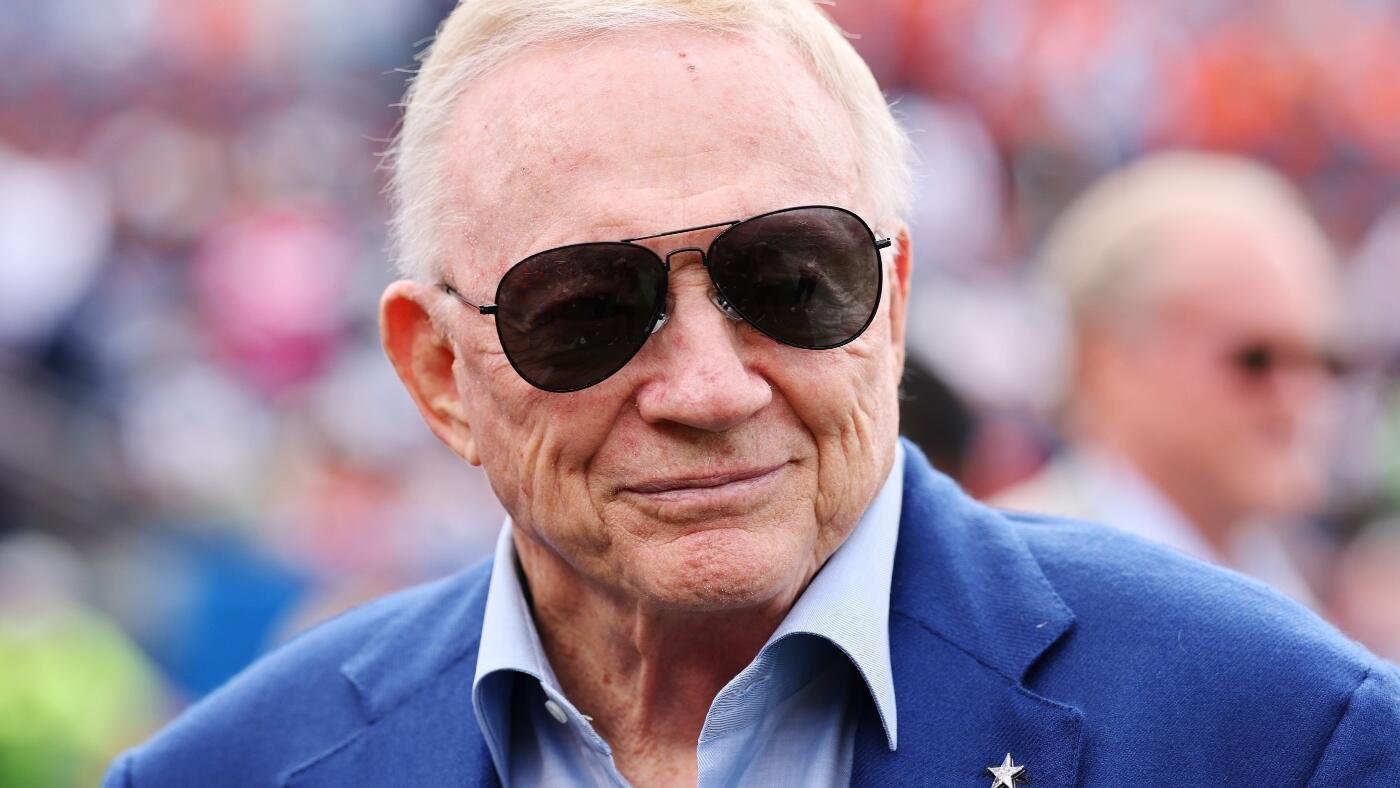 An older man with white hair wears dark aviator sunglasses and a blue blazer over a light blue shirt, standing before a blurred crowd.