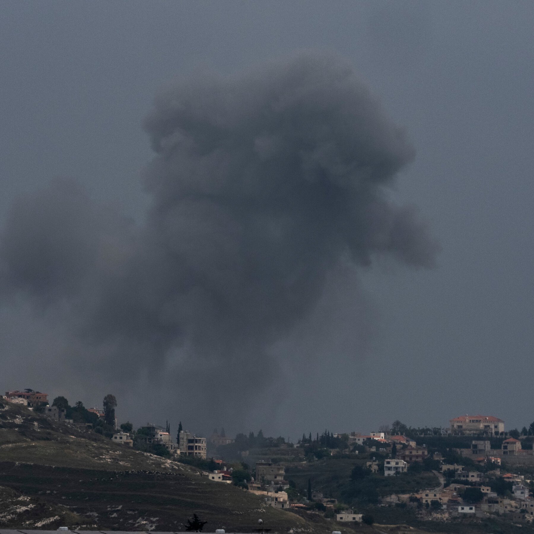 A large plume of dark smoke rises from a hilly landscape scattered with light-colored buildings and trees under a gray sky.