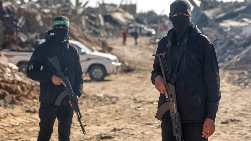 Two masked, armed individuals in dark clothing stand in a debris-filled area with a white pickup truck and damaged structures visible in the background.