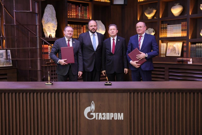 Four men in suits stand behind a wooden podium displaying the "Газпром" (Gazprom) logo, with two holding red folders, in a library-like room with bookshelves.