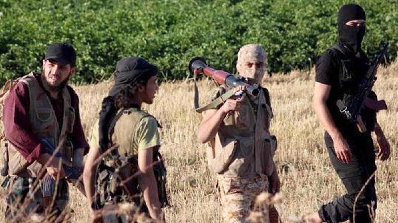 Four armed individuals, some with faces covered, walk through a field carrying rifles and a rocket launcher.