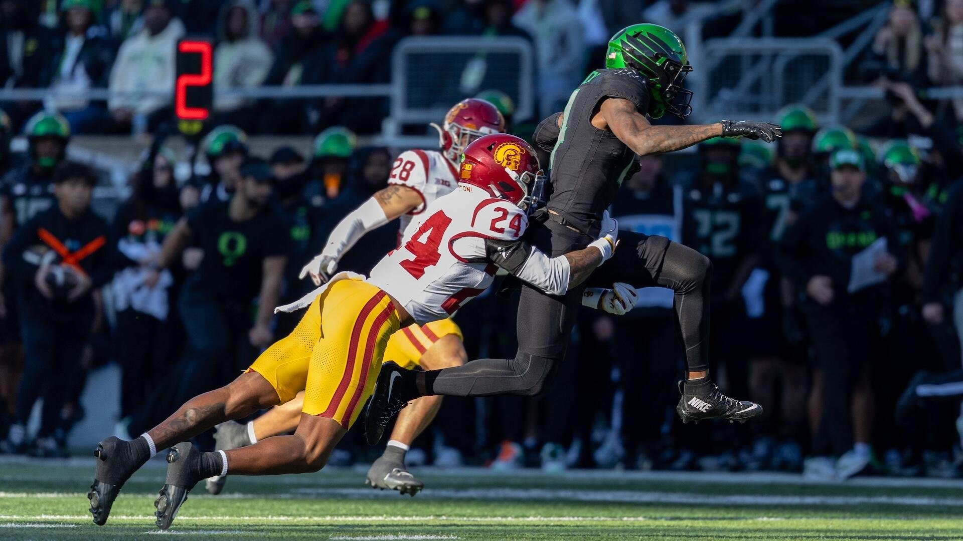 A football player in a white and cardinal #24 jersey tackles a ball carrier in a black uniform with a green helmet during a game.