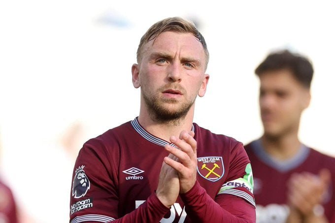 A bearded man in a maroon West Ham United jersey applauds with a serious expression, while another player in matching attire stands blurred behind him.