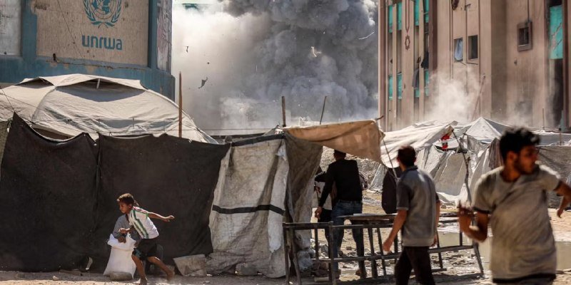 Smoke billows behind UNRWA-branded tents and displaced people walking through a makeshift camp.