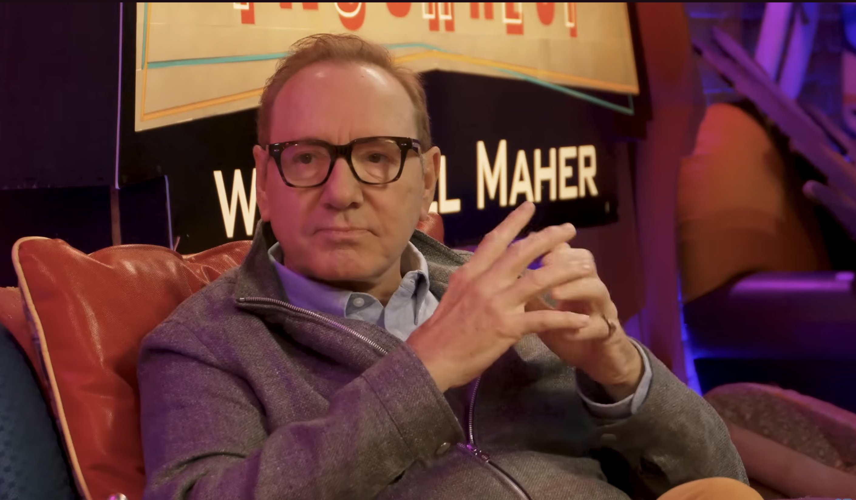 A bespectacled man in a gray zip-up jacket sits gesturing with his hands in front of a sign reading "...AL MAHER."