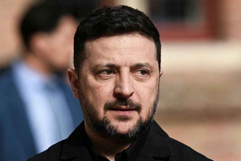 A man with dark hair and a beard, wearing a black shirt, looks off to the side with a serious expression while a blurred figure stands behind him.