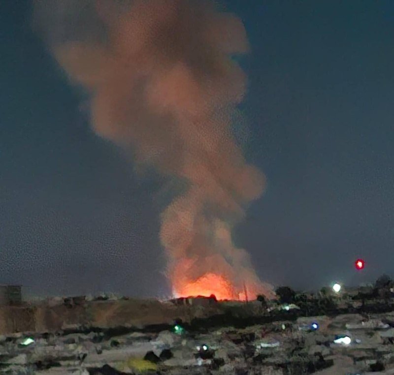 A large fire erupts at night near a settlement, sending a massive plume of orange-lit smoke into the dark sky above buildings.