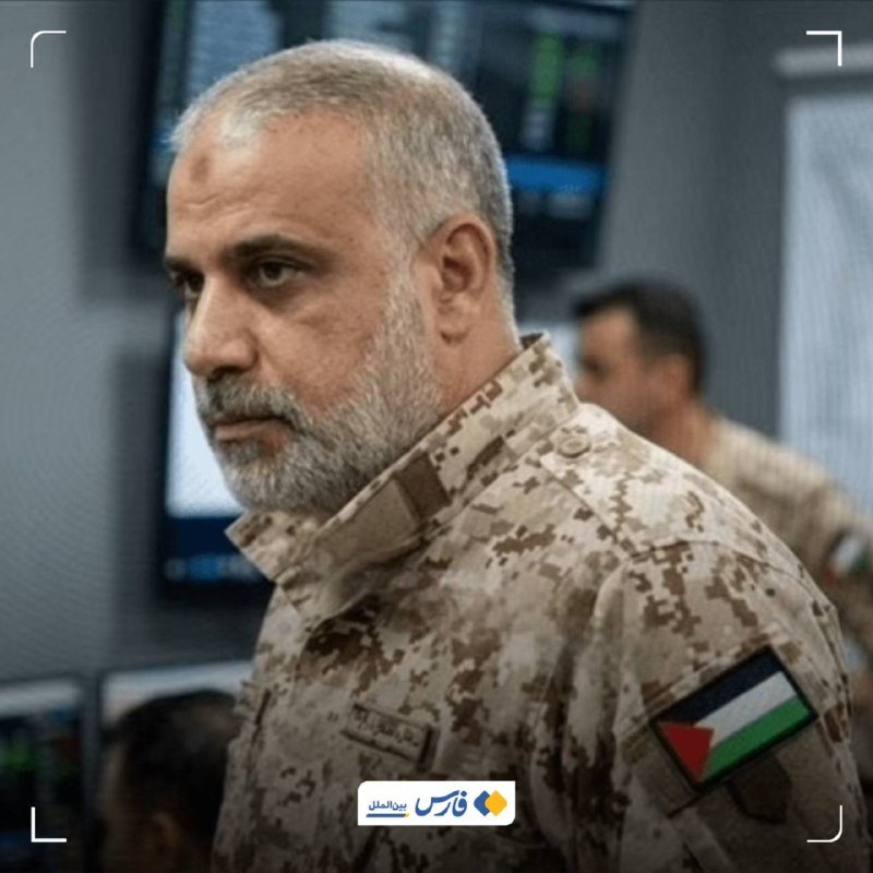 A man with a short beard wearing a desert camouflage uniform with a Palestinian flag patch stands in what appears to be a control room with monitors in the background.