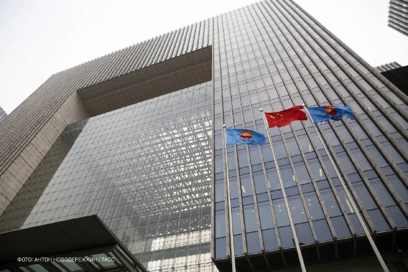 Three flags, including one red with yellow stars, fly in front of a modern glass skyscraper photographed from below, with a photo credit in Cyrillic at the bottom left.