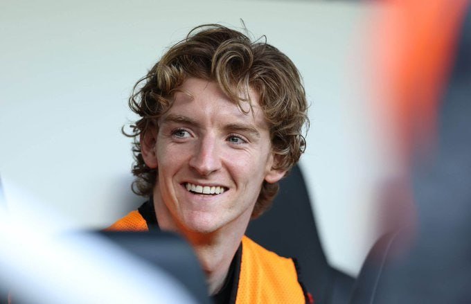 A curly-haired young man in an orange top smiles broadly while looking off-camera.