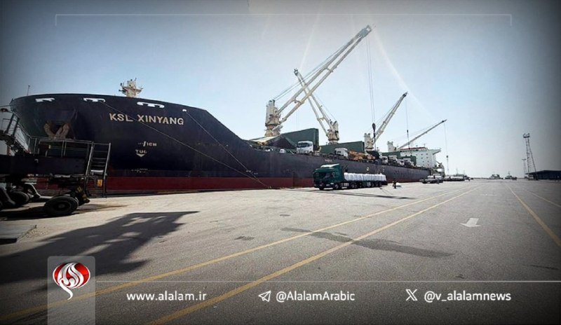 A large cargo ship labeled "KSL XINYANG" is docked at a port, with cranes positioned above it and trucks nearby on the concrete pier.