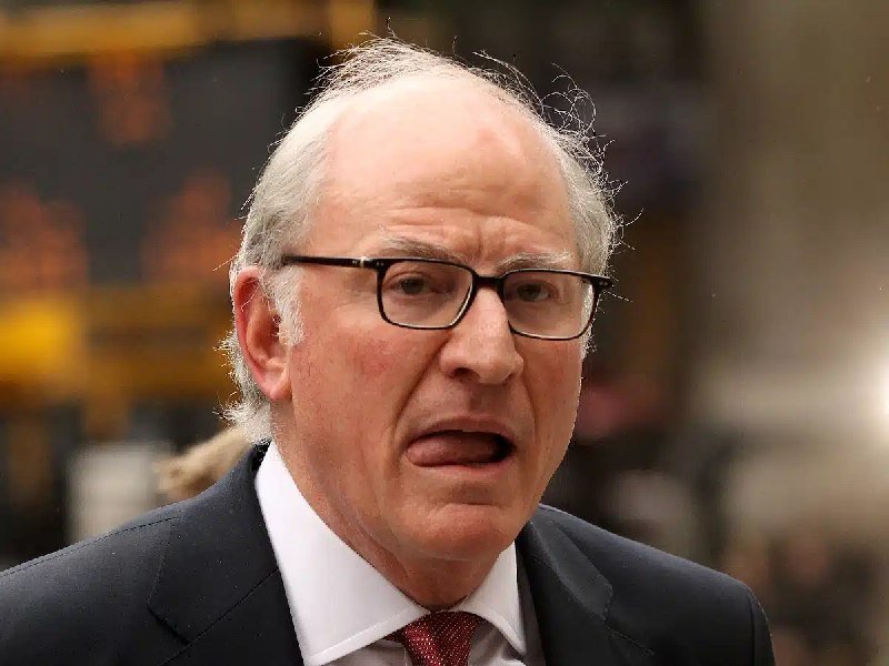 An older man with glasses, wearing a dark suit and red tie, speaks outdoors against a blurred background.
