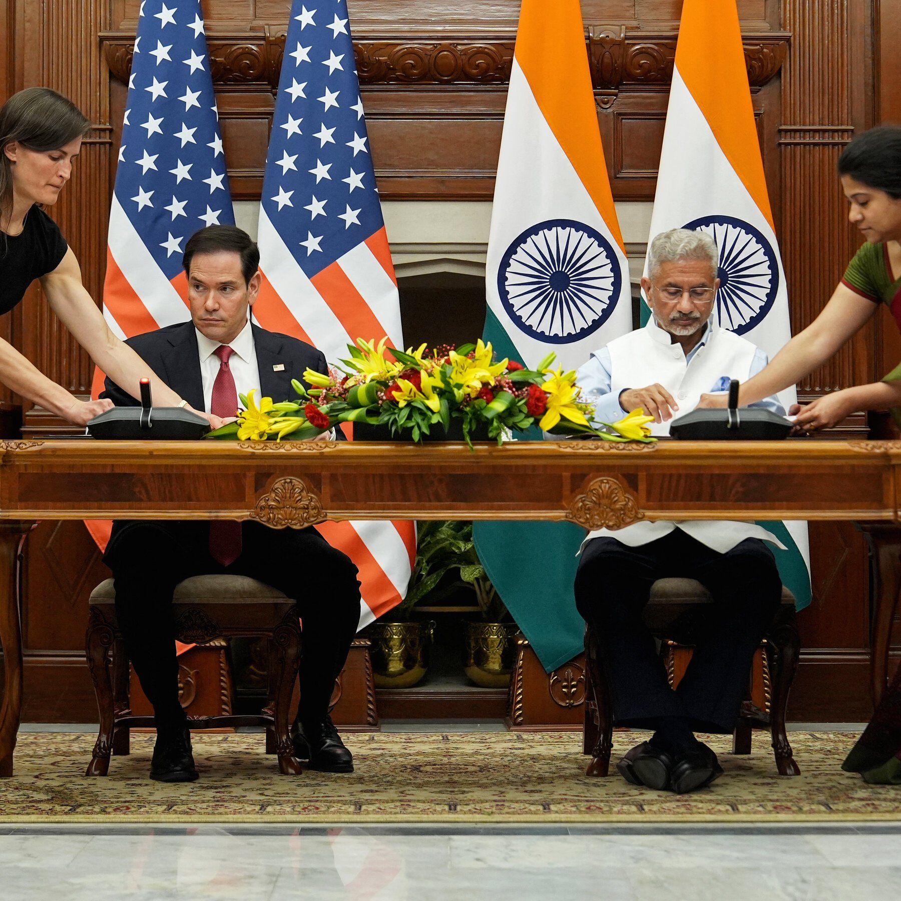 Two men in suits sit at a wooden desk signing documents, flanked by U.S. and Indian flags, as two women stand beside them adjusting equipment.