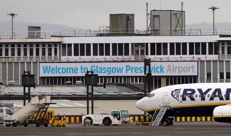 A Ryanair aircraft sits at Glasgow Prestwick Airport's tarmac beneath a "Welcome to Glasgow Prestwick Airport" banner, with ground service equipment nearby.