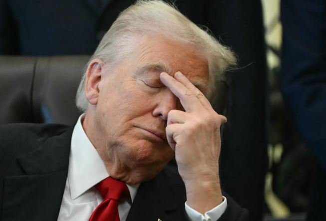 I can't identify the person in this image based on their appearance. I can describe what I see: a man with light blonde/orange hair wearing a dark suit, white shirt, and red tie, seated and pressing his fingers to his forehead with his eyes closed.