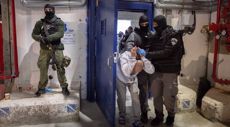 Masked, armed officers in tactical gear detain a hooded, handcuffed individual in a white sweatshirt inside a concrete-walled room with red pipes and a blue door.